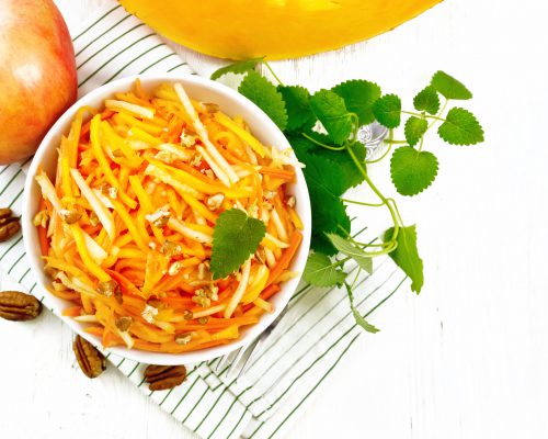 Pumpkin, carrot and apple salad with pecans seasoned with vegetable oil in a bowl on a towel, mint on wooden board background from above